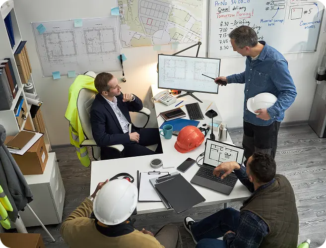 Engineering team in a planning meeting around a desk with blueprints, laptops, and hard hats; one person presents with a pointer at a monitor.