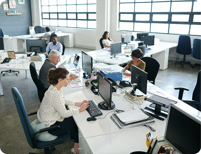 Open-plan office with multiple employees at white desks using computers; woman in the foreground types at a keyboard.