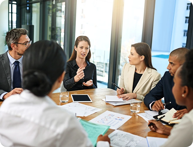 Six professionals in a modern office sit around a wooden table, a woman speaking as others listen.