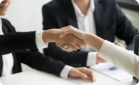 Two professionals in business attire shake hands across a white table, signaling a deal.
