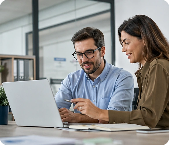 Two colleagues discuss a laptop screen at a shared desk in an office setting.