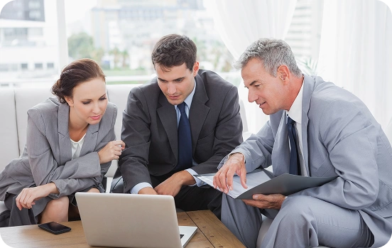Three business professionals, two men and a woman, lean in to review something on a laptop during a meeting in a bright office.