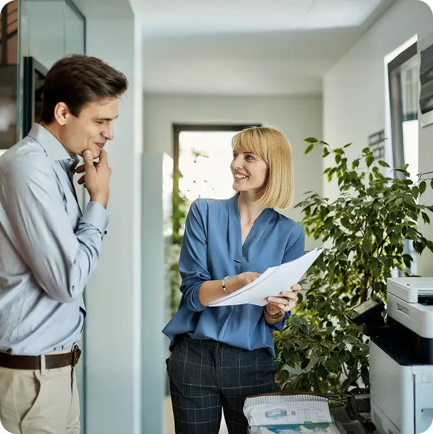 Two coworkers in a bright office; woman in blue blouse showing papers while smiling at a man thoughtfully listening in a hallway.